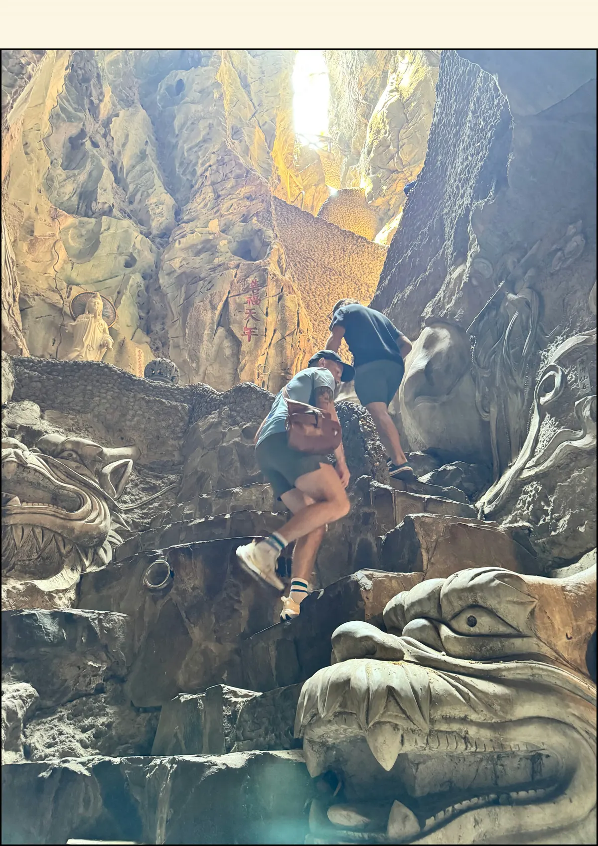 Two people climbing rock steps inside a sunlit cave beside a carved stone dragon