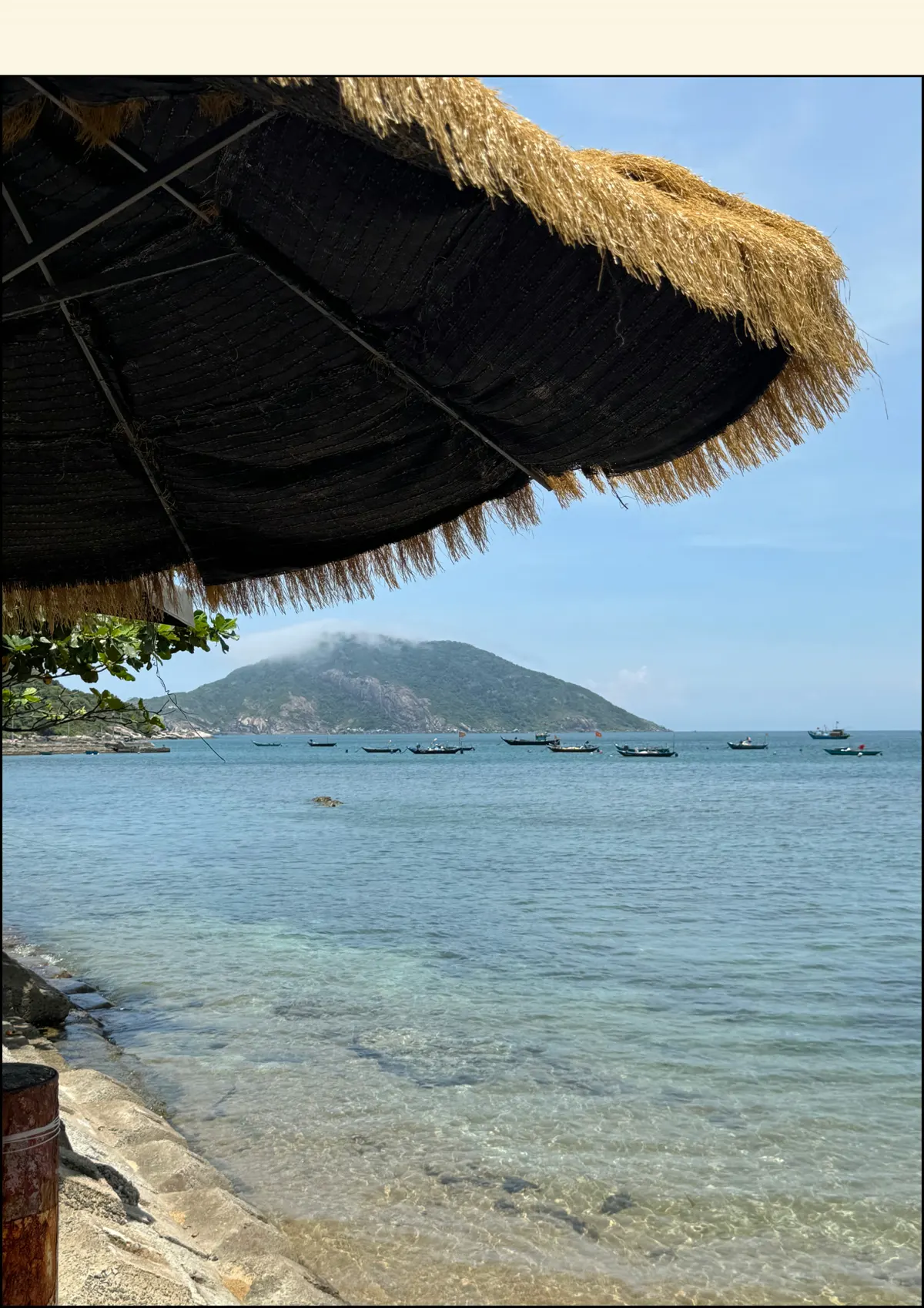 Clear shallow sea and a pale beach seen from beneath a thatched umbrella, with a green island on the horizon
