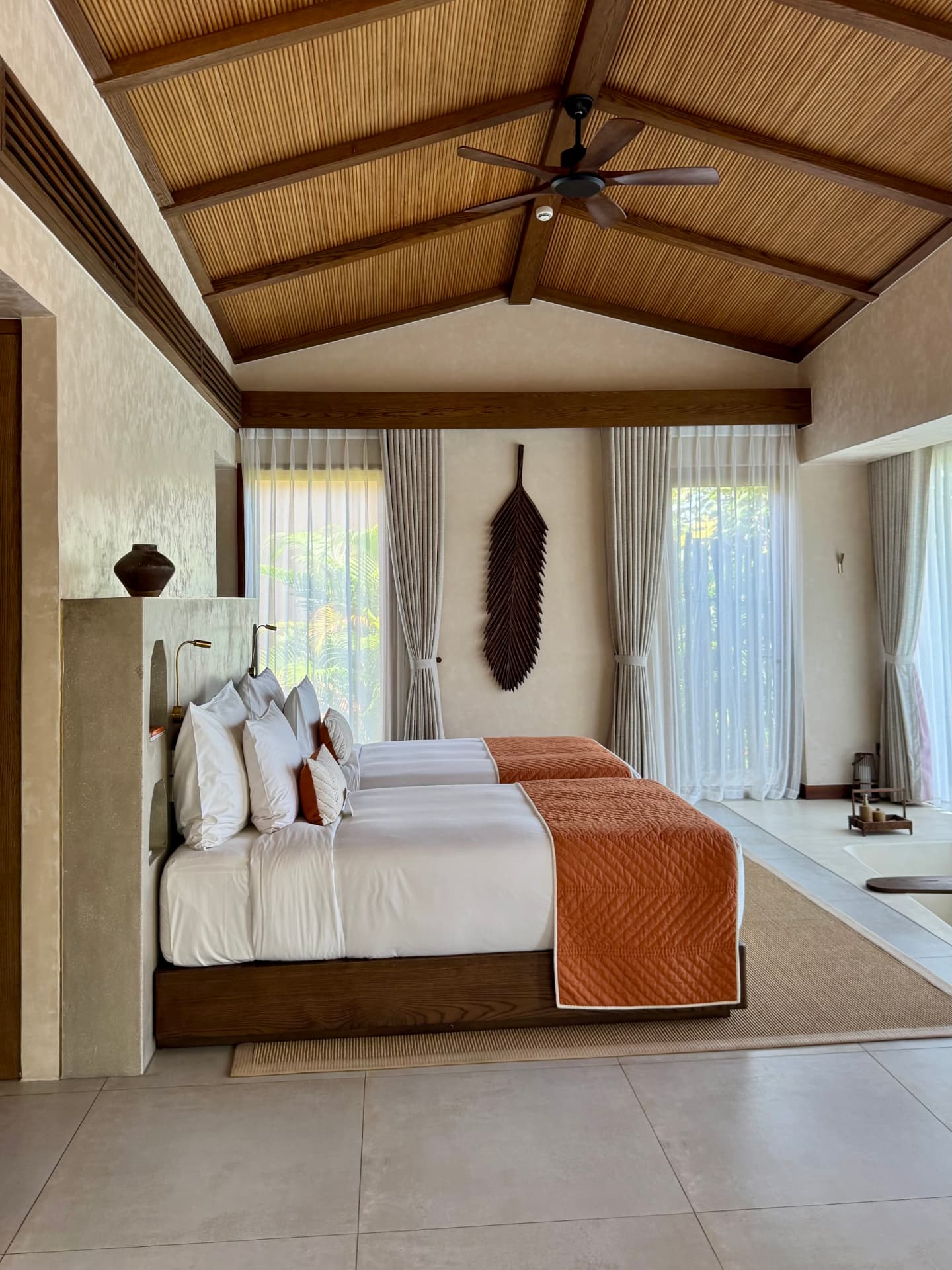The bedroom of a private pool villa, with two timber platform beds, sheer drapes and tall glass doors opening onto tropical planting